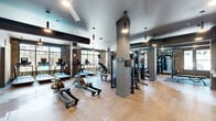 Located at 4090 Rio Vista Dr in Colorado Springs, CO, this contemporary fitness center offers a well-appointed, single-floor layout designed to accommodate a full range of workout routines. The space is divided into distinct functional zones, creating an organized and efficient environment for users of all fitness levels.

The main workout floor features light-toned herringbone hardwood flooring throughout, lending a clean and modern aesthetic. Exposed spiral ductwork runs along the ceilings, complementing the industrial-modern design, while pendant lighting fixtures provide focused illumination across the space. Brick accent walls add warmth and texture, contrasting with the smooth, painted structural columns that define different workout areas.

The cardio zone is well-equipped with treadmills, rowing machines, ellipticals, and a stair climber, all positioned near large windows that invite ample natural light and views of the surrounding outdoor area. A dedicated free weights section includes a full dumbbell rack, flat benches, and rubber-matted flooring, while cable machines and strength equipment occupy an adjacent area lined with floor-to-ceiling mirrors.

A separate cycling studio features a row of stationary bikes, expansive windows overlooking a pool area, and mirrored walls that enhance the sense of space. An additional room provides space for functional training accessories including stability balls, resistance bands, and kettlebells, with a Peloton screen mounted for guided workouts.