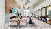 Located at 4555 Grove Springs Cir in Apopka, Florida, this single-floor community clubhouse offers a generously sized, thoughtfully designed gathering space. The grand living room serves as the centerpiece, boasting soaring double-height ceilings, dramatic slatted wood ceiling details, and an abundance of natural light from floor-to-ceiling windows. A terrazzo floor anchors the main lounge area, which features a sleek built-in fireplace, wall-mounted TV, and elegant sculptural chandeliers. Plush seating arrangements invite relaxation alongside curated contemporary artwork. The living space extends into a vibrant game room with a full-size billiards table, slatted wood ceiling accents, built-in banquette seating with textured wall panels, and warm hardwood-style flooring. The dining area is equally refined, with bold geometric blue wallcoverings, framed artwork, and modern bistro-style tables set on light hardwood floors. The dedicated office area features a built-in workstation with pendant lighting set against a soft blue accent wall, complemented by patterned carpet flooring and adjacent cabinetry with open shelving. A connecting hallway provides seamless access between spaces, finished with glass-panel black-framed doors that maintain visual openness throughout the facility.