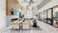 Located at 4555 Grove Springs Cir in Apopka, Florida, this single-floor community clubhouse offers a generously sized, thoughtfully designed gathering space. The grand living room serves as the centerpiece, boasting soaring double-height ceilings, dramatic slatted wood ceiling details, and an abundance of natural light from floor-to-ceiling windows. A terrazzo floor anchors the main lounge area, which features a sleek built-in fireplace, wall-mounted TV, and elegant sculptural chandeliers. Plush seating arrangements invite relaxation alongside curated contemporary artwork. The living space extends into a vibrant game room with a full-size billiards table, slatted wood ceiling accents, built-in banquette seating with textured wall panels, and warm hardwood-style flooring. The dining area is equally refined, with bold geometric blue wallcoverings, framed artwork, and modern bistro-style tables set on light hardwood floors. The dedicated office area features a built-in workstation with pendant lighting set against a soft blue accent wall, complemented by patterned carpet flooring and adjacent cabinetry with open shelving. A connecting hallway provides seamless access between spaces, finished with glass-panel black-framed doors that maintain visual openness throughout the facility.
