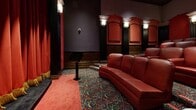 Located at 909 W Temple St in Los Angeles, this elegantly appointed private screening room delivers a true cinematic experience through its rich, theatrical design. The single-floor space is dressed in a deep crimson and dark wood palette that evokes classic Hollywood glamour from the moment you enter through its connecting hallways.

The main screening area is the centerpiece of the space, featuring rows of plush red leather reclining seats arranged in a tiered layout to ensure unobstructed sightlines to the screen. A grand curved stage with dramatic red velvet curtains trimmed in gold anchors the far wall, framing the screen with unmistakable theatrical flair. Flanking the stage are tall, fluted black columns topped with ornate capitals and wall sconces that cast a warm, intimate glow throughout the room.

The walls are finished with dark wood wainscoting and arched decorative panels, adding architectural depth and a refined, formal character. Recessed coffered ceiling panels with embedded spotlights provide layered lighting that enhances the cinematic atmosphere. The floor is covered in a boldly patterned carpet in deep, jewel-toned hues, complemented by red aisle runners that guide guests to their seats. Small accent tables are thoughtfully placed throughout the seating area. The overall ambiance is immersive, luxurious, and unmistakably inspired by the golden age of cinema.