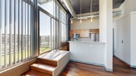 This urban loft on Cuming Street in Omaha's vibrant downtown offers a striking industrial-modern aesthetic across a well-proportioned, single-level floor plan. The open living area immediately impresses with soaring ceilings, exposed ductwork, and floor-to-ceiling windows fitted with vertical blinds that flood the space with abundant natural light and offer sweeping city views. Rich hardwood flooring flows consistently throughout the entire unit, anchoring the open layout with warmth. A raised platform with wooden steps adds a distinctive architectural element near the kitchen, which features warm wood cabinetry, stainless steel appliances including a microwave and refrigerator, a light stone countertop, and pendant Edison bulb lighting overhead. Exposed brick accents in select areas reinforce the building's industrial character. The two bedrooms each benefit from generous window access and ample natural light, with dedicated closets offering wire shelving systems for organized storage. Both bathrooms are finished with warm wood vanity cabinetry, light stone countertops, large mirrors, and overhead cabinet storage, with one bathroom featuring a full soaking tub. A connecting hallway ties the layout together efficiently, completing this well-designed urban retreat.