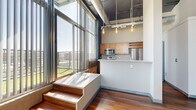 This urban loft on Cuming Street in Omaha's vibrant downtown offers a striking industrial-modern aesthetic across a well-proportioned, single-level floor plan. The open living area immediately impresses with soaring ceilings, exposed ductwork, and floor-to-ceiling windows fitted with vertical blinds that flood the space with abundant natural light and offer sweeping city views. Rich hardwood flooring flows consistently throughout the entire unit, anchoring the open layout with warmth. A raised platform with wooden steps adds a distinctive architectural element near the kitchen, which features warm wood cabinetry, stainless steel appliances including a microwave and refrigerator, a light stone countertop, and pendant Edison bulb lighting overhead. Exposed brick accents in select areas reinforce the building's industrial character. The two bedrooms each benefit from generous window access and ample natural light, with dedicated closets offering wire shelving systems for organized storage. Both bathrooms are finished with warm wood vanity cabinetry, light stone countertops, large mirrors, and overhead cabinet storage, with one bathroom featuring a full soaking tub. A connecting hallway ties the layout together efficiently, completing this well-designed urban retreat.