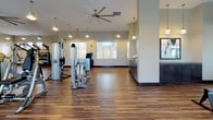 This expansive single-floor fitness center is thoughtfully designed to accommodate a comprehensive range of workout needs. The space features rich, dark hardwood-style flooring throughout, complemented by warm neutral-toned walls and large windows that allow generous natural light to fill the interior while providing outdoor views of the surrounding community.

The cardio zone lines one side of the facility, housing a row of treadmills, ellipticals, and stationary bikes, all positioned to face the windows—offering an airy, open feel during workouts. Large wall-mounted mirrors extend along one section, visually expanding the space and supporting proper form during exercise. A wall-mounted flat-screen television is centrally positioned for entertainment.

The strength training area is well-appointed with Cybex cable machines and multi-functional weight systems strategically arranged across the floor. A dedicated free weights section includes a full dumbbell rack, kettlebells, medicine balls, and a functional training rig with a punching bag, supporting a variety of training styles. Open floor space in this zone accommodates stretching and floor exercises.

Overhead, large ceiling fans with pendant lighting fixtures provide comfortable air circulation. A service counter with dark cabinetry offers convenient access to towels and amenities, rounding out this fully equipped fitness facility.