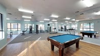 This single-floor amenity space seamlessly blends fitness and recreation into one open, well-lit environment. The room is bright and airy, benefiting from multiple windows that invite natural light to pour across the warm hardwood flooring. A row of fluorescent ceiling fixtures ensures consistent illumination throughout.

The fitness area is thoughtfully equipped with a cable weight machine, treadmills, and additional cardio and strength-training equipment, catering to a range of workout routines. A wall-mounted television is positioned for convenient viewing during exercise. Full-length mirrors line one wall, visually expanding the space while providing functional feedback for users during their workouts.

Complementing the fitness zone, the recreation area features a classic billiards table with a rich wood frame and a vibrant blue felt surface, serving as a natural focal point of the room. The combination of fitness and leisure amenities makes this space well-suited for both active exercise and social enjoyment. The light blue walls contribute to a clean, calm ambiance, and the open layout provides ample room to move freely between activity areas.