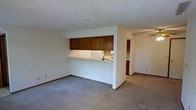 This single-floor space is designed with an open and functional layout that connects the living area to the kitchen through a convenient pass-through counter. The living area is finished with neutral gray carpeting that extends throughout, providing a soft, cohesive foundation. White walls and standard-height ceilings keep the space feeling clean and bright.

The kitchen is partially open to the living area, separated by a raised counter that serves as both a visual divider and a casual dining or prep surface. Dark wood cabinetry lines the upper portion of the kitchen wall, offering generous storage space. White appliances, including a refrigerator and range, are visible within the kitchen, complementing the light-colored countertops and neutral palette.

Beyond the main living area, a hallway leads to additional rooms, with a ceiling fan visible in the adjoining space, suggesting a bedroom or secondary living area. Dark wood cabinetry is also visible further down the hall, indicating continued kitchen or utility storage. The overall layout is practical and straightforward, making efficient use of the available space across a single floor.