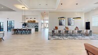 Located at 1055 Winnall Ln in Fayetteville, NC, One at Fox Mile's clubhouse is a thoughtfully designed, single-floor amenity space that spans a generous footprint. The interior greets visitors with a soaring entry foyer featuring dramatic vaulted ceilings, an eye-catching starburst chandelier, bold patterned accent walls, and light wood-look flooring that flows throughout the entire space.

The main living area is anchored by floor-to-ceiling arched windows that bathe the room in natural light and offer views of the outdoor pool. Elegant ring chandeliers hang above plush white seating arrangements, creating a refined yet welcoming lounge atmosphere. A second living area showcases a striking navy geometric tile accent wall with floating shelves, complemented by a sage green wet bar with pendant lighting.

A long communal table with upholstered chairs serves as a central co-working hub, while a dedicated coffee bar with bold encaustic tile backsplash adds a café-style touch. A leasing or business desk area with patterned wallcovering provides a professional workspace zone.

The game room features a herringbone wood-panel accent wall, a pool table, large-screen TVs, and designer pendant lighting. Glass double doors open to a fully equipped fitness center, completing this dynamic, amenity-rich community gathering space.