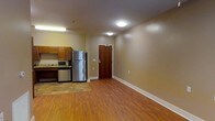 This single-floor unit offers an open and functional layout, combining the kitchen and living area into one cohesive space. The kitchen is equipped with stainless steel appliances, including a full-sized refrigerator and microwave, complemented by warm-toned wooden cabinetry and a tiled floor section that visually distinguishes it from the main living area. The remainder of the space features durable hardwood-style flooring in a rich, warm tone that extends throughout, adding a sense of continuity and warmth to the interior. The walls are finished in a neutral beige tone, contributing to a clean and inviting ambiance. Ceiling-mounted flush lighting provides consistent illumination across the space. The entryway is defined by a solid wood-paneled door with a modern electronic keypad lock, offering both practicality and security. White baseboards provide a crisp contrast against the flooring and walls. The overall design is straightforward and practical, with a layout that maximizes usable floor space while maintaining a comfortable and livable atmosphere throughout.