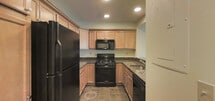 This single-floor space showcases an open-concept design that seamlessly connects the living area and kitchen. The living space features wall-to-wall neutral carpeting, recessed lighting, and crown molding that adds a refined architectural detail along the ceiling. A floor vent provides climate control, and the neutral-toned walls create a clean, versatile backdrop throughout.

The kitchen is defined by a raised breakfast bar with a dark granite-style countertop, offering both a functional workspace and a natural divider between the cooking and living areas. Light wood cabinetry lines the kitchen walls, providing ample storage, while a black refrigerator anchors the space. Recessed lighting above the kitchen ensures a well-lit work environment.

Beyond the living area, a hallway leads to the entry door, suggesting additional rooms or storage accessible from this corridor. The floor transitions from carpet to a hard surface near the entryway, adding practical durability in higher-traffic zones. Overall, the space presents a functional and cohesive layout with a neutral, move-in-ready aesthetic.