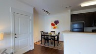 This single-floor space showcases a thoughtfully designed dining area that flows naturally into the kitchen. The dining space features rich, dark hardwood flooring grounded by a patterned area rug, creating a warm and inviting atmosphere. A dark-stained dining table with four matching chairs serves as the focal point of the area, complemented by a vibrant floral artwork on the crisp white walls that adds a pop of color to the neutral palette. Modern track lighting mounted on the ceiling provides focused illumination, enhancing both functionality and style. The kitchen is partially visible, revealing light stone countertops atop white cabinetry, seamlessly integrating with the open-concept layout. White panel doors with clean, classic molding contribute to the space's polished aesthetic. The overall design balances contemporary elements with warm, traditional touches, resulting in a cohesive and comfortable living environment. The white walls and bright ceiling work together to maximize the sense of openness throughout the space.