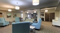 This single-floor communal space offers an open, welcoming layout designed for both relaxation and social interaction. The main lounge area is anchored by plush light blue sofas and accent chairs arranged around small side tables, creating inviting conversation clusters. Cylindrical pendant lights hang at varying heights throughout the space, providing warm, even illumination across the patterned carpeted floors. Structural columns clad in stacked stone add a natural, textural element that contrasts with the smooth neutral-toned walls and ceiling. A raised ceiling detail with recessed lighting defines a transitional zone, visually separating the lounge from an adjoining area that features darker cabinetry and countertops, suggesting a kitchenette or café-style bar. Beyond this, additional seating arrangements and windows allow natural light to filter into the space. Artwork on the walls adds touches of color and character throughout. The overall design blends contemporary and transitional styles, with a cohesive color palette of blues, creams, and warm wood tones, creating a comfortable and functional communal environment.