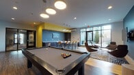 Located at 480 Rantoul Street in Beverly, Massachusetts, this single-floor community amenity space is thoughtfully designed to offer residents a dynamic and versatile gathering environment. The space is anchored by a central game room featuring a full-sized pool table beneath a cluster of modern pendant lighting, creating a lively focal point for social activity. Light wood flooring flows throughout, complemented by bold accent walls in steel blue and vibrant yellow that lend the space a contemporary, energetic character. A dedicated lounge area furnished with sleek leather chairs and a wall-mounted flat-screen TV provides an intimate setting for relaxing or watching entertainment. Adjacent to the game room, a well-appointed kitchenette includes dark cabinetry, a stainless steel microwave and refrigerator, a sink, and ample counter space — all oriented toward a communal high-top dining table with bar seating for a crowd. Large floor-to-ceiling windows and glass doors bathe the space in natural light and offer views of an adjacent outdoor area, seamlessly connecting the interior to the exterior. The overall ambiance is modern and inviting, blending functional amenities with stylish design details suited for both casual gatherings and community events.