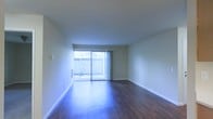 This single-floor space presents a clean, open layout highlighted by rich, dark hardwood flooring that extends throughout the main living area. The floors are well-maintained and add a sense of warmth and depth to the room. Smooth, neutral-toned walls complement the flooring and contribute to a bright, airy atmosphere. White baseboards provide a crisp contrast along the perimeter of the room.

A prominent sliding glass door serves as a central architectural feature, drawing in generous amounts of natural light and offering direct access to an outdoor balcony or patio area. The exterior space appears to include a railing, suggesting an elevated position with outward views. Sheer white curtains frame the door, allowing light to diffuse softly into the interior.

The ceiling is flat and uniform, maintaining a straightforward aesthetic throughout the space. The open floor plan provides flexibility for a variety of furniture arrangements, making the living area feel functional and adaptable. Multiple electrical outlets are visible along the walls, reflecting practical design considerations. Overall, the space conveys a simple, well-maintained environment suited for comfortable everyday living.