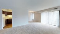This single-floor space presents a clean, understated aesthetic defined by neutral tones and an open, airy layout. The living area is generously proportioned, featuring soft, plush wall-to-wall carpeting in a warm beige tone that adds comfort underfoot and contributes to the overall sense of calm throughout the space. Smooth white ceilings reflect natural light effectively, enhancing the brightness of the interior. Large windows dressed with vertical blinds allow for adjustable natural light, flooding the rooms with a soft, diffused glow while maintaining privacy. The walls are finished in a warm off-white tone, providing a versatile backdrop well-suited to a variety of furnishing styles. A centrally positioned flush-mount ceiling light fixture provides ambient illumination, complementing the natural light from the windows. The layout flows openly between areas, with a structural beam subtly defining the transition between spaces without interrupting the overall sense of openness. The interior is move-in ready, offering a blank canvas that is both functional and adaptable. Overall, the space exudes a simple, practical elegance with an emphasis on light, space, and comfort.