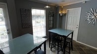 This single-floor space presents a thoughtfully designed interior with a bold, contemporary aesthetic. The dining area is a standout feature, showcasing dark charcoal-gray walls that create a dramatic yet inviting atmosphere. Two bar-height tables with sleek frosted glass tops are paired with matching black stools, offering a modern and functional dining arrangement.

Natural light filters into the room through large windows dressed with horizontal blinds, balancing the deep color palette with a bright, airy feel. Overhead, a pendant light with an industrial-style fixture provides focused illumination, complementing the wall-mounted sconce nearby that adds a warm, ambient glow.

Decorative elements elevate the space with intentional character. A large typographic wall canvas featuring an inspirational "Life" quote serves as a bold focal point, while an ornate arched mirror with a geometric design adds an elegant contrast to the otherwise modern setting. Crown molding along the ceiling adds a refined architectural detail, lending a polished finish to the room.

The flooring features large-format stone-look tiles in neutral tones, grounding the space and providing a durable, easy-to-maintain surface. A white paneled door offers a clean contrast against the dark walls. Overall, the space combines functionality with a strong design identity, making it both practical and visually compelling.