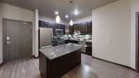 This 2-bedroom, 2-bathroom residence is designed with a modern aesthetic and functional layout across a single floor. The kitchen is a standout feature, showcasing rich dark cabinetry, granite countertops, a central island with pendant lighting, stainless steel appliances, and a decorative tile backsplash — all set atop wood-style flooring that extends into the open living area. The living space is generously sized, filled with natural light, and accented by a chandelier and ceiling fan, creating a warm and inviting atmosphere. The primary bedroom offers a comfortable retreat with plush carpeting, a ceiling fan, and windows overlooking greenery, accompanied by a private en-suite bathroom featuring a dual-sink vanity with granite countertops, wood-style flooring, and a walk-in closet with wire shelving. The second bedroom is equally well-sized with soft carpeting and neutral tones. The second bathroom includes a full soaking tub and shower combination with brushed nickel fixtures and wood-style flooring. A dedicated closet and an in-unit stacked washer and dryer add exceptional convenience. Throughout, dark wood trim and door accents provide a cohesive, contemporary finish.