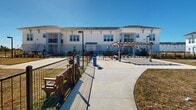 This dog park in Wesley Chapel, FL, offers a well-designed outdoor space for canine companions and their owners. The park is characterized by expansive grassy areas, divided into separate sections by metal fencing, allowing for safe off-leash play. Paved walkways wind through the park, providing easy access and comfortable strolling paths for visitors.

The park is surrounded by modern, multi-story residential buildings, creating a cohesive community atmosphere. These structures, painted in light, neutral tones, feature balconies and ample windows, suggesting a pet-friendly living environment. The architecture blends harmoniously with the park's layout, enhancing the overall aesthetic appeal.

Amenities within the park include shaded areas, courtesy of a wooden pergola, offering respite from the Florida sun. Bench seating is strategically placed, allowing owners to relax while supervising their pets. Lamp posts are installed throughout, ensuring the area is well-lit for evening use.

The landscaping is minimalist yet functional, with newly planted trees dotting the perimeter, promising future shade and greenery. The open layout of the park allows for excellent visibility and a sense of spaciousness, making it an ideal spot for both energetic play and leisurely dog walks.