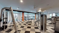 Located at 3000 Hicks St in Houston's vibrant 77007 corridor, this single-floor amenity facility is designed to deliver a comprehensive fitness and social experience. The space is anchored by a spacious main gym area with a striking black-and-white checkered floor, housing a wide array of strength and cardio equipment including cable machines, a squat rack, free weights, and dumbbell racks. Large floor-to-ceiling windows flood the workout areas with natural light and offer views of the outdoor surroundings, creating an open and energizing atmosphere. Ceiling fans and recessed lighting maintain comfort throughout.

A dedicated spin and studio room features hardwood-style flooring, a wall-mounted flat-screen TV, mirrored walls, and a row of stationary bikes, complemented by a large window overlooking a landscaped courtyard. The lounge and social area showcases bold design with vibrant orange accent walls, a geometric multi-colored mural, and contemporary black furniture arranged around a marble-top coffee table with a wall-mounted TV. High-top tables provide additional casual seating. Black-framed glass doors connect the zones, reinforcing the modern aesthetic that defines this dynamic, well-appointed community fitness facility.