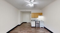Located at 1410 SW Broadway in Portland, Oregon, this efficiently designed studio unit offers a practical and functional layout across a single floor. The living area is finished with durable wood-style flooring and crisp white walls, creating a clean and bright atmosphere. A ceiling fan with integrated lighting provides both airflow and illumination throughout the open space. Crown molding adds a subtle architectural detail, while a classic cast-iron radiator offers reliable heating. A multi-pane window in the living area invites natural light and frames views of the surrounding greenery. The kitchenette is neatly arranged along one wall, featuring warm-toned wood cabinetry, a stainless steel sink with a countertop, a white electric range, and a compact stainless steel refrigerator. A textured ceiling adds a traditional character to the space. The bathroom is thoughtfully appointed with wood-style flooring that continues from the main area, a wall-mounted sink, a toilet, and a bathtub with a shower rod — offering all essential amenities in a compact footprint. The overall layout makes efficient use of every square foot, making this unit a practical option in the heart of Portland.