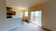 This single-floor space offers an open-concept layout that seamlessly connects the living area, dining space, and kitchen. The living area is finished with plush carpet flooring, providing a comfortable and inviting atmosphere, while the adjoining dining and kitchen zones transition to durable hardwood-style flooring. Warm beige walls throughout create a cohesive and welcoming ambiance.

A sliding glass door leads to a private outdoor balcony, allowing generous natural light to fill the interior and offering views of the surrounding neighborhood. Vertical blinds on the sliding door provide convenient light control and privacy. A secondary window near the dining area further brightens the space.

The dining area is highlighted by an elegant chandelier pendant light, adding a touch of refinement to the open layout. The kitchen features rich, dark wood cabinetry and a light-toned countertop with a raised bar-style partition that defines the space without fully closing it off from the rest of the unit.

A wall-mounted HVAC unit ensures year-round climate comfort. Clean white trim and baseboards provide a crisp contrast to the warm wall tones, completing the well-maintained and move-in-ready presentation of this thoughtfully designed space.
