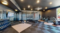 This contemporary fitness center is thoughtfully designed to support a wide range of workout routines in a sleek, modern environment. The space is anchored by a striking functional training rig stocked with a full selection of kettlebells, medicine balls, battle ropes, and resistance bands, set against a boldly patterned feature wall that adds visual depth to the room. A heavy punching bag and flat bench further complement the strength and conditioning zone.

The cardio section is positioned along a wall of floor-to-ceiling windows, flooding the area with natural light and offering open outdoor views. Equipment includes spin bikes, elliptical trainers, a rowing machine, and treadmills, providing ample options for cardiovascular training. A dedicated dumbbell rack with a full weight range rounds out the free weights offerings.

Underfoot, a geometric checkerboard-patterned carpet tile floor in contrasting tan and charcoal tones adds a dynamic visual element while providing a durable workout surface. The dark ceiling with recessed and pendant lighting creates a focused, energetic atmosphere throughout. A ceiling fan aids in air circulation over the main training floor.

Practical amenities include a dual-level water fountain set into a textured hexagonal tile wall near the glass entry doors, and generous open floor space allowing for stretching and bodyweight exercises. The overall design balances functionality with a polished, modern aesthetic.