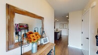 This single-floor home is thoughtfully designed to balance functionality and comfort throughout its well-appointed layout. A charming entryway welcomes visitors with warm hardwood-style flooring, a rustic wooden console table, and a large wood-framed mirror, setting the tone for the home's inviting aesthetic. The hallway leads seamlessly into the open-concept kitchen and living area, where dark espresso cabinetry, granite-style countertops, and stainless steel appliances anchor the kitchen. A breakfast bar with industrial-style bar stools provides casual seating and connects the kitchen to the adjacent living space. The living room features vaulted ceilings, plush carpeting, and ample room for relaxed gatherings. A standout feature of the home is a dedicated hobby or collector's room, showcasing extensive built-in shelving and display cases. The carpeted bedrooms offer comfortable retreats with natural light from large windows. Two bathrooms are finished with dark wood vanities and hardwood-style flooring. A practical in-unit laundry room houses a full-size washer and dryer alongside the home's utility systems. Throughout, neutral wall tones and consistent flooring materials create a cohesive and welcoming atmosphere.