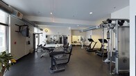 This fitness center offers a thoughtfully designed workout space built to accommodate a full range of exercise needs. The room features a dark, rubberized floor that provides both durability and traction, complemented by clean white walls and a smooth white ceiling with recessed lighting that keeps the space bright and well-illuminated. Exposed ventilation ductwork runs along the ceiling, lending an industrial aesthetic to the otherwise sleek interior.

Large mirrors line the walls, visually expanding the space and allowing users to monitor their form during workouts. The cardio section is well-stocked with treadmills and additional aerobic machines, while the strength training area includes a cable machine, a rowing machine, and a variety of weight equipment, including a dumbbell rack. Natural light filters in through a window on one side of the room, adding to the overall brightness of the environment.

The layout is open and spacious, allowing for easy movement between equipment stations without overcrowding. The overall ambiance is clean, functional, and professional, making this fitness center a practical and inviting space for a comprehensive workout routine.