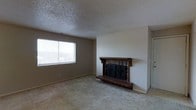 This single-floor space presents a straightforward and functional layout centered around a welcoming living area. The room's most distinctive feature is a classic brick fireplace accented by a dark wood mantel and ornate iron glass doors, serving as a natural focal point. A raised brick hearth adds to its traditional charm and provides a cozy atmosphere. The living area is finished with wall-to-wall carpeting in a neutral tone, contributing to a warm and comfortable feel underfoot. A wide window allows natural light to filter into the space, offering views to the outside and brightening the otherwise neutral interior. The walls are painted in a soft, understated hue that complements the overall palette. Overhead, a textured popcorn ceiling spans the room, consistent with classic residential construction. The open layout of the living area provides flexibility for a variety of furniture arrangements. The space combines traditional architectural details with practical finishes, making it a versatile and livable environment suitable for everyday use.