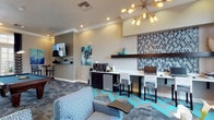 This thoughtfully designed single-floor amenity space blends functionality and contemporary style throughout its interconnected areas. The interior is unified by a cohesive color palette of teal, navy, and warm neutrals, accented by patterned upholstery and bold geometric flooring that adds visual energy underfoot.

The lounge area features plush, patterned armchairs arranged around a dark wood coffee table, framed by large windows dressed with navy drapes that invite natural light. Abstract artwork adds a curated, modern touch to the walls. A striking sputnik-style chandelier serves as a focal point overhead, complementing recessed lighting and crown molding details throughout.

Adjacent to the lounge, a dedicated business center offers a long countertop workstation with multiple computer stations, a tiled accent wall, floating shelves, and a coffee/beverage setup with a mini-refrigerator beneath—ideal for productivity.

The game room is highlighted by a full-sized billiards table set beneath pendant lighting, surrounded by French doors that flood the room with natural light and provide access to adjoining spaces. High-top bistro tables with bar seating offer additional casual gathering spots, completing this well-appointed communal amenity space.