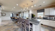Located at 2090 Columns Dr in Cumming, GA, this single-floor communal kitchen and dining space is thoughtfully designed to offer both functionality and comfort. The open layout seamlessly integrates the kitchen and dining areas, creating an inviting environment for gathering and socializing.

The kitchen is a standout feature, anchored by a substantial white brick-clad island topped with a dark stone countertop and surrounded by upholstered bar-height seating. Elegant pendant lights hang above, providing focused task lighting with a stylish touch. White cabinetry lines the perimeter, complemented by a glossy subway tile backsplash, dark stone countertops, and stainless steel appliances including a full-size refrigerator. Recessed lighting and crown molding add a polished finish throughout.

The dining area accommodates multiple round wooden tables with metal chairs and blue cushioned seating, arranged beneath recessed lighting and crown molding. Wainscoting along the lower walls adds a classic architectural detail, while large windows dressed with Roman shades and floor-length drapes allow abundant natural light to fill the space. A fireplace with a decorative mantle serves as a focal point in the adjoining area. Multicolored slate tile flooring runs throughout, tying the entire space together with a warm, earthy aesthetic. French doors connect to additional rooms, enhancing the open and accessible feel of the layout.