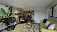 This single-floor space showcases a distinctive, nature-inspired interior design that blends organic elements with modern aesthetics. The living area serves as the visual centerpiece, anchored by a dramatic accent wall clad in realistic birch tree panels, accompanied by a bold green "ADVENTURE" lettering that adds a playful, expressive touch. Dark hardwood floors run throughout, providing a grounding contrast to the lighter furnishings and decor.

The living space is furnished with black rattan-style chairs, a chartreuse sofa, and a standout live-edge wood coffee table on hairpin legs, adding an artisanal, natural quality to the room. A geometric green-and-cream area rug ties the seating arrangement together with a retro-inspired pattern.

The layout flows openly into the dining area, which is distinguished by a decorative wall treatment featuring an asymmetric arrangement of square panels in neutral and green tones, alongside a pendant light above a round dining table with modern chairs. Together, the spaces create a cohesive, eclectic ambiance that balances nature-inspired elements with a contemporary, design-forward sensibility.