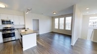 This well-appointed single-floor residence offers a thoughtfully designed open-concept layout that seamlessly connects the kitchen and living areas. The kitchen features crisp white cabinetry, a dark countertop island with a built-in sink, and a track lighting fixture overhead, providing both functionality and a clean, contemporary aesthetic. Dark hardwood-style flooring runs throughout the space, offering a rich contrast against the light, neutral-toned walls and bright white trim.

The living area is generously proportioned, benefiting from a trio of large windows dressed with white horizontal blinds that invite an abundance of natural light while maintaining privacy. A flush ceiling light fixture adds warmth to the space. A doorway visible from the main living area leads to an additional room, suggesting a flexible layout suited for various living arrangements.

The overall ambiance is modern and move-in ready, characterized by clean lines, a neutral color palette, and quality finishes throughout. The combination of open living space, practical kitchen design, and ample natural light makes this a functional and inviting single-floor residence.