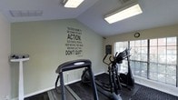 This single-floor fitness center is thoughtfully designed to provide residents or members with a functional and motivating workout environment. The space features a clean, open layout with durable dark flooring that defines the exercise area and provides a stable surface for cardio equipment, including treadmills and an elliptical cross-trainer. A large motivational quote by Conrad Hilton is prominently displayed on the soft green accent wall, serving as an inspiring focal point for those working out. The angled ceiling adds an architectural dimension to the room, while overhead fluorescent lighting ensures the space is well-lit at all times. A window fitted with blinds allows natural light to filter in and provides a view of the surrounding greenery, contributing to an airy and pleasant atmosphere. A wall clock keeps exercisers mindful of their time, and additional informational signage is visible near the window. The overall ambiance is clean, practical, and motivating — a dedicated space designed to encourage consistent fitness activity.