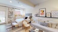 Located at 2003 Greene St in Columbia, South Carolina's vibrant 29205 zip code, this single-floor studio offers a thoughtfully designed open-concept layout that seamlessly combines sleeping, lounging, and living areas. The main space is defined by warm neutral tones, recessed lighting, and a striking exposed brick accent wall that adds rustic character alongside the otherwise clean, modern aesthetic. The sleeping area features a generous bed with a gray wood-panel headboard, while the living area accommodates a slipcovered sofa and rattan seating centered around a glass-top coffee table. A picture ledge shelf displays curated artwork, contributing to the space's stylish, curated feel. A full-length ornate white mirror reflects light throughout the room, enhancing the sense of openness. The compact kitchenette is efficiently outfitted with dark cabinetry, white countertops, a stainless steel undermount sink with a matte black faucet, an induction cooktop, and an over-the-range microwave. The full bathroom features large-format dark gray tile flooring, crisp white walls, a dark vanity with a white countertop, and a shower with a decorative curtain — clean and functional in design. Natural light filters in through a window overlooking the exterior, completing this well-appointed urban studio.