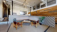 Located at 1710 E Broad St in Richmond, Virginia, this thoughtfully designed residential lobby showcases an impressive blend of industrial history and contemporary aesthetics. The space is anchored by a striking, large-scale preserved Armour Extract of Beef mural painted directly on the original brick wall, serving as a vivid reminder of the building's storied past. Soaring ceilings amplify the sense of openness, while an eye-catching geometric chandelier with brass-toned arms and pendant lights adds a modern, artistic focal point overhead. Upper-level clerestory windows with industrial-style steel frames allow natural light to filter in, complementing the floor-to-ceiling glass entry doors that offer views of East Broad Street. A welcoming lounge area features an L-shaped upholstered banquette backed by patterned encaustic-style tilework, paired with leather-and-wood safari chairs arranged around a rustic wood coffee table. Reclaimed wood wall paneling adds warmth and texture throughout. Practical amenities include a traditional mailbox bank, a smart package locker system, an elevator with decorative tropical-print wallcovering, and a staircase with wood treads and a metal railing. The polished concrete floors tie the industrial character of the space together seamlessly.