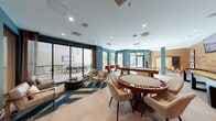 This single-floor amenity lounge is thoughtfully designed as a multi-zone social and recreation space. The interior is defined by bold teal accent walls, warm wood-paneled feature walls, and large-format light tile flooring that unifies the open layout. Recessed lighting and modern pendant fixtures illuminate the space throughout.

The gaming area is a central highlight, equipped with a full-size pool table in rich walnut tones, a dedicated shuffleboard table, and a round poker table with built-in chip holders surrounded by upholstered seating. Dual flat-screen TVs are mounted on the wood-paneled wall, creating an entertainment focal point. A cork accent wall with decorative artwork adds textural contrast nearby.

A comfortable lounge zone features plush armchairs, a round coffee table, and a bold patterned area rug. Large sliding glass doors connect this area seamlessly to an outdoor patio furnished with dining tables and chairs and an additional mounted TV. Natural light flows generously through the floor-to-ceiling glazing, enhancing the open, airy atmosphere. High bar-height seating with red chairs provides additional social gathering spots, completing this well-appointed communal space.