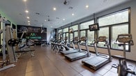 This single-floor fitness center is thoughtfully designed to support a comprehensive workout experience. The space is flooded with natural light through an expansive wall of large, dark-framed windows that frame views of lush greenery, creating an energizing and inviting atmosphere throughout. Durable rubber tile flooring covers the entire facility, providing both safety and comfort underfoot.

The cardio zone lines the windowed perimeter and features a generous row of treadmills, each equipped with individual flat-screen monitors, alongside stair climbers and elliptical machines similarly outfitted with personal screens. Ceiling-mounted televisions provide additional entertainment options for longer sessions.

The strength training area is equally well-appointed, offering a variety of cable machines and multi-functional weight stations arranged to allow efficient use of the space. A dedicated free weights section includes a full dumbbell rack, while stability accessories such as medicine balls of varying weights and exercise balls are neatly organized on dedicated racks.

An accent wall featuring bold fitness-themed graphic artwork adds a motivational focal point to the space. Recessed lighting supplements the abundant natural light, ensuring the facility remains well-lit at all times. A secondary area near the entrance provides additional cardio equipment along with a frosted-glass privacy partition and direct access to an adjoining outdoor area.