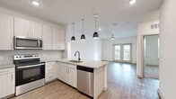 This well-designed 1-bedroom, 1-bathroom residence offers a thoughtful layout across a single floor, combining modern finishes with functional living spaces. The open-concept kitchen and living area serves as the heart of the home, featuring a central island with granite countertops and a built-in sink, complemented by three sleek pendant lights overhead. White shaker-style cabinetry lines the walls, while a full suite of stainless steel appliances — including a refrigerator with ice maker, electric range, microwave, and dishwasher — adds a polished, contemporary touch. Light hardwood-style flooring flows throughout the main living areas, and French doors at the far end of the living room invite natural light and provide access to an outdoor space. A ceiling fan overhead adds comfort to the airy living area. The bedroom is a comfortable retreat finished with soft carpeting, a ceiling fan, and a window with blinds. It connects directly to the full bathroom, which features a granite-topped vanity, a combined tub and shower with white tile surround, and wood-style flooring. A dedicated walk-in closet with wire shelving provides ample storage. Clean white walls and recessed lighting throughout contribute to a bright, modern ambiance.