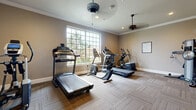 This fitness room offers a dedicated space for cardiovascular exercise, thoughtfully designed with both function and comfort in mind. The room is finished with neutral warm-toned walls and crisp white crown molding that runs along the perimeter, lending a polished, clean aesthetic throughout. Durable carpet tile flooring in a subtle herringbone pattern covers the entire floor, providing a practical and cushioned surface ideal for a workout environment. Two ceiling fans keep the space well-ventilated, complemented by recessed lighting that ensures the room is well-lit at all times. A large multi-pane window on one wall floods the space with natural light and offers a pleasant outdoor view of surrounding greenery, creating an inviting atmosphere during workouts. The room is equipped with multiple treadmills, a stationary bike, and an elliptical trainer, catering to a variety of cardio fitness needs. Along the walls, a low bench provides a convenient spot for stretching or equipment storage, while informational fitness charts, a wall clock, and a sanitizing station add practical touches. The space is accessed through a classic paneled door and benefits from a security camera, ensuring a safe and organized environment. Overall, this fitness room strikes a balance between a functional workout facility and a comfortable, well-appointed space.