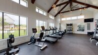 This well-appointed fitness facility is housed within a single-story structure designed in a modern farmhouse architectural style. The interior is immediately distinguished by its dramatic vaulted ceiling, where rich, dark-stained exposed wood beams intersect against white-paneled shiplap walls, creating a striking visual contrast. Large-format pendant lights and an oversized ceiling fan hang from the peak of the ceiling, contributing both functional lighting and a refined aesthetic.

Natural light is a defining feature of the space, with large windows lining the lower walls and a row of clerestory windows positioned near the roofline, allowing daylight to pour in from multiple angles. Views of the surrounding outdoor amenities and landscaping are visible through the windows, further enhancing the open, airy atmosphere.

The rubber-floored exercise area is divided into distinct functional zones. The cardio section is equipped with Life Fitness treadmills, ellipticals, a recumbent bike, and a rowing machine. A dedicated strength-training zone features a cable machine, weight racks loaded with free weights, and a bench press station. Full-length mirrors line one wall to assist with form, while a wall-mounted television provides entertainment. A water fountain is conveniently located near the exit.
