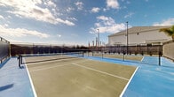 Located in Kissimmee, Florida, this outdoor recreational facility features multiple dedicated pickleball courts designed to accommodate players of all skill levels. The courts are finished in a striking combination of blue and green surfacing, with crisp white boundary lines clearly delineating each playing area. Sturdy nets are installed at regulation height on each court, ensuring a game-ready experience at all times.

The entire facility is enclosed by a robust chain-link perimeter fence, providing a secure and defined playing environment. Wind screens line portions of the fencing, helping to reduce wind interference during play. Tall light poles are positioned throughout the complex, enabling evening and nighttime use of the courts.

The surrounding landscape features palm trees and manicured greenery, reflecting the characteristic Florida setting. Neighboring residential buildings are visible beyond the fence line, situating this facility within a community context. The open-air design takes full advantage of the Florida sunshine, with the broad, unobstructed sky contributing to a bright and energetic atmosphere. Whether for casual play or organized matches, this well-maintained pickleball complex offers a high-quality outdoor sporting experience in the heart of Kissimmee.