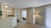 This single-floor residential space is designed with a practical and comfortable layout that maximizes flow between its key living areas. The open-concept living room is finished with soft wall-to-wall carpeting in neutral beige tones, creating a warm and inviting atmosphere. A floor lamp in the corner adds a touch of warmth and highlights the clean, simple lines of the space.

The living area transitions seamlessly into a kitchen that features a raised breakfast bar countertop in white, offering both a casual dining surface and a visual partition between the two spaces. The kitchen is equipped with a range and overhead exhaust hood, with cabinetry providing functional storage. Recessed lighting in the kitchen adds a clean, modern touch.

A doorway from the living area leads directly into a private bedroom, which is visible from the main room and features its own carpeted flooring consistent with the rest of the unit. The bedroom provides a quiet, enclosed retreat. The space benefits from a ceiling with a textured finish and is well-ventilated with visible HVAC vents throughout. Neutral wall colors and understated finishes give the entire space a cohesive and move-in-ready appeal.