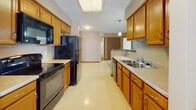 This single-floor residence at 2116 Deer Lakes Dr in Buffalo, New York offers a well-organized layout designed for comfortable everyday living. The interior is finished with neutral beige tones throughout, creating a cohesive and understated aesthetic across all rooms.

The galley-style kitchen is a functional highlight, featuring warm honey-toned wood cabinetry with ample upper and lower storage, light-colored countertops, a double-basin sink, and black appliances including a refrigerator, range, built-in microwave, and dishwasher. A bright skylight panel and pendant lighting illuminate the space, while durable speckled vinyl flooring adds practicality. In-unit laundry hookups are also visible toward the end of the kitchen area.

The living and bedroom areas are carpeted throughout, providing warmth underfoot. A ceiling fan with integrated lighting serves one of the rooms, alongside a window fitted with blinds that allows natural light to filter in. The entry area features a solid front door beside a framed window, creating a welcoming foyer.

Additional features include a large double-door closet, a bathroom with cabinetry, and multiple connected rooms visible in the three-dimensional floor plan scan. Recessed vents, smoke detectors, and consistent wood baseboards and door trim detail the space throughout.
