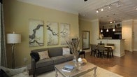 This thoughtfully designed single-floor space exudes a warm, sophisticated ambiance through its cohesive use of neutral tones and carefully curated details. The living area serves as the heart of the home, anchored by a plush charcoal sofa and a sleek metallic coffee table that adds a contemporary touch. A striking triptych of nature-inspired artwork lines the soft olive-toned walls, lending an organic, artistic character to the room. Track lighting overhead provides adjustable illumination, complementing the overall warm atmosphere. Rich hardwood flooring flows seamlessly from the living area into the dining space, creating a sense of continuity and openness. The dining area accommodates a full table and chair set, with pendant lighting suspended above to define the space and add an intimate feel. Just beyond, a glimpse of the kitchen reveals pendant lights and a clean, well-appointed layout. Decorative accents, including a tall dried botanical arrangement and a framed mirror, add layers of texture and depth throughout. The open-concept layout encourages a natural flow between living, dining, and kitchen areas, making the space feel both functional and inviting — ideal for everyday living and entertaining alike.