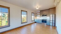 This well-designed one-bedroom, one-bathroom unit offers an efficient and thoughtfully laid-out single-floor living space. The open-concept kitchen and living area serves as the heart of the unit, featuring light wood flooring that runs throughout, crisp white walls, and multiple grid-pane windows with warm wood trim that flood the space with natural light and offer pleasant outdoor views of the surrounding neighborhood. The kitchen is outfitted with two-tone cabinetry — taupe upper cabinets paired with light blue-gray lower cabinets — stainless steel appliances including a range and built-in microwave, open wood shelving, and a classic white subway tile backsplash. The bedroom is a cozy, private retreat with its own window providing natural light and outdoor views, a wall-mounted mini-split HVAC unit for individualized climate control, and a sliding-door closet offering convenient storage. The bathroom features a taupe vanity with a white countertop and undermount sink, tile flooring, and a clean, accessible layout with grab bar hardware. A hallway connects the rooms seamlessly, completing this efficiently planned and move-in-ready living space with modern, neutral finishes throughout.