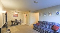 This single-floor space presents an open-concept layout that connects the living and dining areas into one cohesive, functional zone. The living area is carpeted and accommodates a plaid sofa accented with a decorative pillow, while a wall-mounted flat-screen television anchors the adjacent dining space, which features a dark wood table and matching chairs. A ceiling fan with integrated lighting is centrally positioned, providing both airflow and ambient illumination throughout the shared space. The walls are painted in a neutral beige tone, complemented by textured ceilings that add subtle visual character. Framed artwork decorates the walls, contributing a personalized touch to the interior. Toward the back of the space, a set of bifold closet doors and a standard interior door suggest convenient storage options and access to additional rooms. A thermostat is visibly mounted on the wall, indicating individual climate control. The layout is straightforward and efficient, with a clear sightline from the living area through to the hallway, making the overall space feel open and easy to navigate.