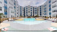 Located at 400 South Greene Street in Greenville, NC, this outdoor amenity courtyard is a standout feature of a modern multi-story residential community. The centerpiece is a generously sized, freeform resort-style swimming pool with a distinctive yellow sun shelf entry zone, surrounded by an expansive pool deck finished in speckled grey and terracotta-toned concrete. Rows of lounge chairs line the perimeter, offering ample space for sunbathing and relaxation.

Adjacent to the main pool, a circular hot tub sits beneath a striking curved black pergola, providing a shaded retreat with lush landscaping and manicured plantings nearby. A dedicated lawn area with ground-level cushions offers an informal lounge space, while a grill station and pergola structure cater to outdoor dining and social gatherings.

Covered lounge areas along the building's ground-floor perimeter feature ceiling fans, comfortable sectional seating, and large glass garage-style doors that seamlessly connect indoor and outdoor spaces. The entire courtyard is framed by the community's bold blue-and-white multi-story façade, accented with palm trees that enhance the resort-inspired atmosphere. This well-appointed outdoor space provides a comprehensive amenity experience centered on leisure and community living.