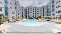 Located at 400 South Greene Street in Greenville, NC, this outdoor amenity courtyard is a standout feature of a modern multi-story residential community. The centerpiece is a generously sized, freeform resort-style swimming pool with a distinctive yellow sun shelf entry zone, surrounded by an expansive pool deck finished in speckled grey and terracotta-toned concrete. Rows of lounge chairs line the perimeter, offering ample space for sunbathing and relaxation.

Adjacent to the main pool, a circular hot tub sits beneath a striking curved black pergola, providing a shaded retreat with lush landscaping and manicured plantings nearby. A dedicated lawn area with ground-level cushions offers an informal lounge space, while a grill station and pergola structure cater to outdoor dining and social gatherings.

Covered lounge areas along the building's ground-floor perimeter feature ceiling fans, comfortable sectional seating, and large glass garage-style doors that seamlessly connect indoor and outdoor spaces. The entire courtyard is framed by the community's bold blue-and-white multi-story façade, accented with palm trees that enhance the resort-inspired atmosphere. This well-appointed outdoor space provides a comprehensive amenity experience centered on leisure and community living.