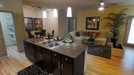 This single-floor residence showcases a thoughtfully designed open-concept layout that seamlessly connects the kitchen, dining, and living areas into one cohesive living space. The kitchen serves as the centerpiece, anchored by a large granite-topped island with a double stainless steel sink, dark espresso cabinetry with brushed nickel hardware, and pendant lighting that casts a warm glow over the workspace. Track lighting overhead ensures the area is well-lit and functional. The kitchen flows naturally into a dining nook and a comfortable living room, where a ceiling fan with a light fixture adds both style and airflow. The living area is appointed with a plush sectional sofa, decorative accent pillows, and a glass-top coffee table, all set atop light hardwood flooring that brightens the space considerably. Warm yellow-toned walls paired with framed artwork lend the interior a welcoming and curated ambiance. A glimpse of a hallway or additional room beyond hints at further private quarters. The overall design balances modern finishes with cozy, residential warmth, making it a functional and aesthetically appealing single-level home.