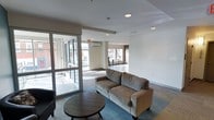 This single-floor space presents a clean, modern aesthetic with an open-plan layout designed to encourage both relaxation and social interaction. The lounge area is anchored by a plush, neutral-toned sofa positioned atop a blue area rug, complemented by a round dark wood coffee table that adds warmth to the contemporary surroundings. Recessed lighting throughout the ceiling provides consistent illumination, while large floor-to-ceiling windows along the perimeter flood the interior with generous natural light and offer views of the surrounding exterior. A wall-mounted split HVAC unit ensures year-round climate control. The flooring transitions between carpeted and hard surface areas, subtly delineating distinct zones within the open space. Toward the rear, a secondary area is visible, furnished with tall bar-height seating and additional windows, suggesting a casual dining or café-style nook. The white walls and neutral finishes contribute to a bright, airy atmosphere throughout. Overall, the space balances functional design with a welcoming ambiance suitable for a variety of uses.