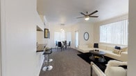 This single-story space showcases an open-concept design that integrates the kitchen, dining, and living areas into one cohesive, flowing environment. The layout is anchored by a dark granite breakfast bar extending from the kitchen, accompanied by sleek adjustable bar stools, making it a functional and stylish transition point between cooking and entertaining spaces.

The dining area is positioned centrally, illuminated by an elegant pendant chandelier that draws the eye upward toward the white ceiling. A ceiling fan with a brushed nickel finish serves the living area, ensuring year-round comfort. The space is finished with plush dark carpeting throughout, providing a rich contrast against the crisp white walls.

The living area is generously proportioned, offering ample room for comfortable seating arrangements. Multiple windows fitted with vertical blinds allow abundant natural light to fill the space while maintaining privacy. A decorative circular mirror accents one wall, adding depth and visual interest. The overall ambiance is clean, contemporary, and versatile, with a neutral black-and-white palette that lends itself well to a variety of interior styling preferences.